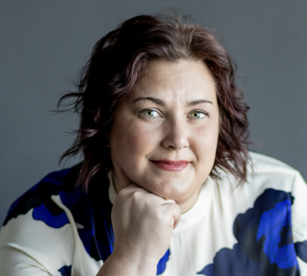 a person with medium length curled brown hair has their fist under their chin as they pose for a headshot